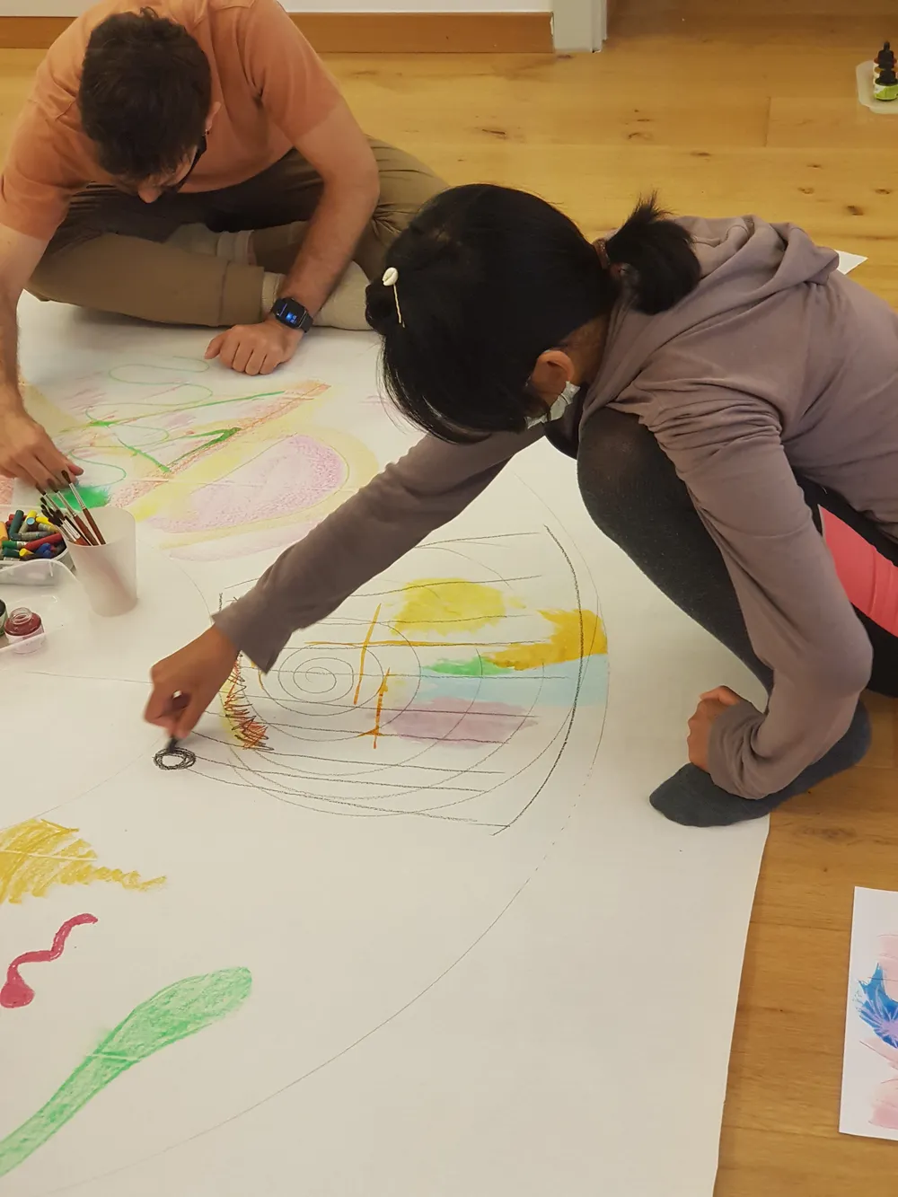 Dues persones adultes dibuixant amb llapis de colors sobre un gran paper estès a terra